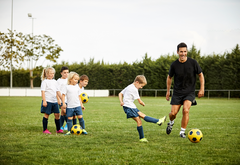 A man and children joyfully playing soccer on a grassy field under a clear blue sky.