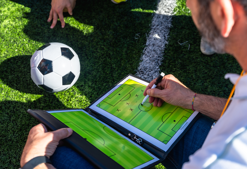 A man sits on a soccer field, writing on a tablet, surrounded by green grass and goalposts in the background.